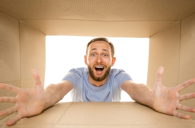 Free photo young astonished man opening the biggest postal package isolated on white. shocked male model on top of cardboard box