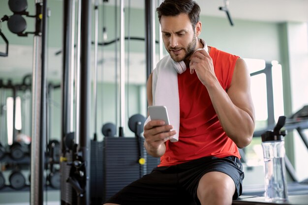 Молодой спортсмен обменивается текстовыми сообщениями на смартфоне во время отдыха в тренажерном зале