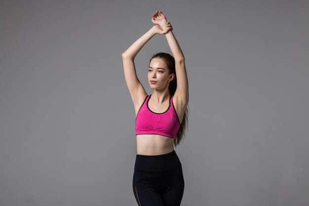 Young athletic fitness woman in sportswear stretching her arms and looking at camera while standing isolated on grey. Healthy lifestyle and sports concept