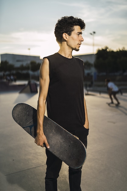 Giovane maschio atletico con un vestito nero che tiene uno skateboard con la mano in tasca