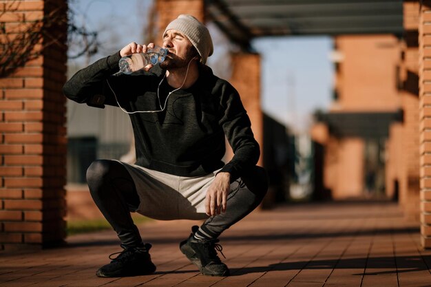 Молодой спортивный мужчина приседает на улице и пьет пресную воду из бутылки.
