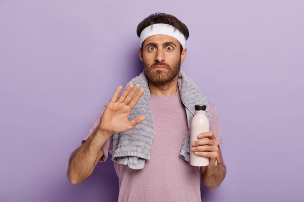 Young athletic man makes refusal gesture, asks not bother him, dressed in active wear, white headband, towel around neck, holds bottle of fresh water