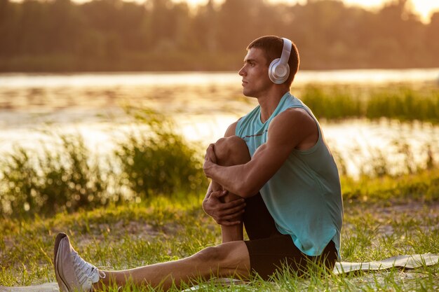 Молодой спортивный мужчина тренируется, слушая музыку на берегу реки на открытом воздухе