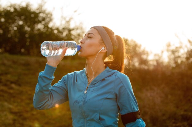 自然の中で朝のランニング中に断水しながら目を閉じて水を飲む若い運動女性