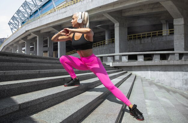 A young athletic woman in shirt and white headphones working out listening to the music at the street outdoors. Doing lunges up stairs. Concept of healthy lifestyle, sport, activity, weight loss.