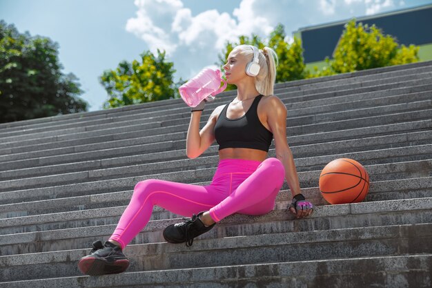 A young athletic woman in shirt and white headphones working out listening to the music at the street outdoors. Sitting on stairs with ball. Concept of healthy lifestyle, sport, activity, weight loss.