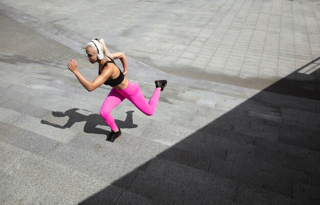 A young athletic woman in shirt and white headphones working out listening to the music at the street outdoors.