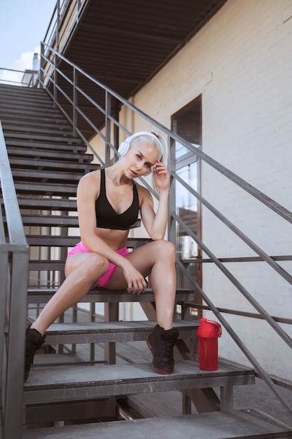 Free photo a young athletic woman in white headphones working out listening to the music on a stairs outdoors. sitting and resting after the training. concept of healthy lifestyle, sport, activity, weight loss.