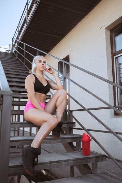 A young athletic woman in white headphones working out listening to the music on a stairs outdoors. Sitting and resting after the training. Concept of healthy lifestyle, sport, activity, weight loss.