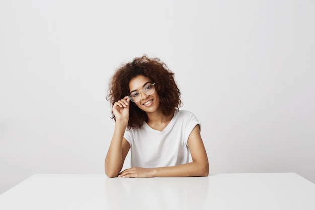 Young attractive african girl in glasses smiling  sitting at the table over white wall. Future fashion icon or graphic designer.