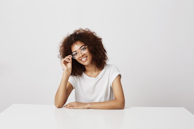 Young attractive african girl in glasses smiling sitting at table over white wall