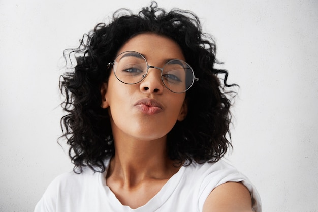 Foto gratuita giovane donna africana attraente che posa con il bacio sulle sue labbra, indossando occhiali alla moda, avendo un aspetto innamorato sentirsi sicuri e belli. affascinante donna dalla pelle scura con i capelli afro divertirsi al chiuso