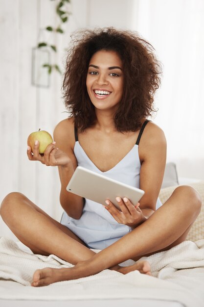 パジャマやパジャマを着て朝の持株タブレットとアップルを笑顔のベッドの上に座っている若い魅力的なアフリカ女性。