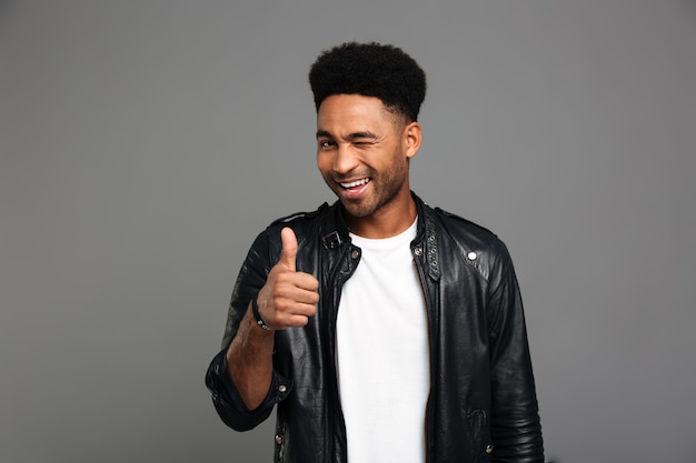 Young attractive afro american boy in leather jacket winks one eye while showing thumb up gesture
