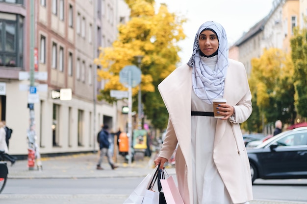 Молодая привлекательная арабка в хиджабе идет по городской улице с сумками и кофе