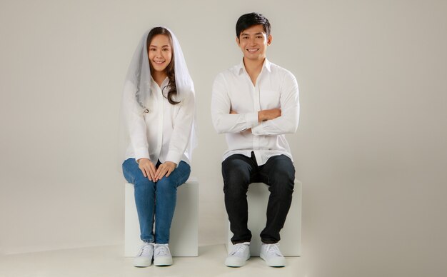 흰 셔츠와 베일을 끼고 손을 잡고 앉아 웃고 있는 젊은 매력적인 아시아 부부