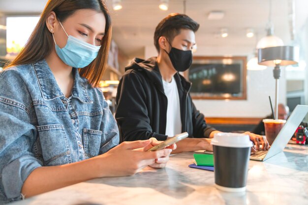 Young attractive asian female and male wearing face mask virus protecting social distancing seat working with laptop in coffee shop cafe