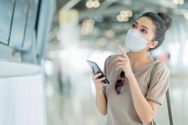 Young attractive asian female traveller wearing face mask hand use smartphone checking map and flight schedule waking with luggage and handbag at airport terminal new normal travel lifestyle