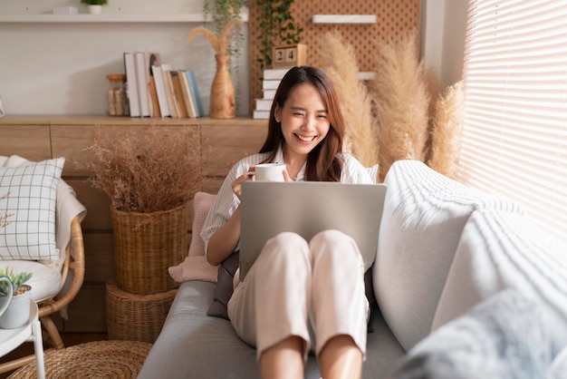 自宅で働く若い魅力的なアジアの女性は、ラップトップチェック検索スケジュール作業プロセス計画レポートを使用してレジャーで朝の光の平和な瞬間とリビングルームのソファでリラックス