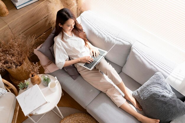 Young attractive asian female working at home hand use laptop checking search schedule working process planning report with leisure relax on sofa in living room with morning light peaceful moment