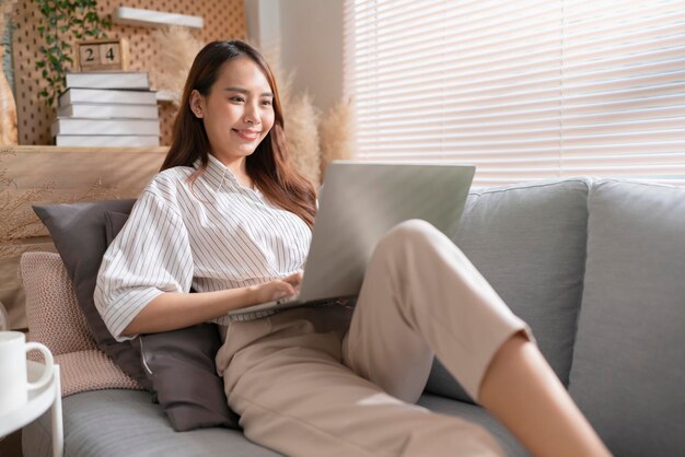 Young attractive asian female working at home hand use laptop checking search schedule working process planning report with leisure relax on sofa in living room with morning light peaceful moment