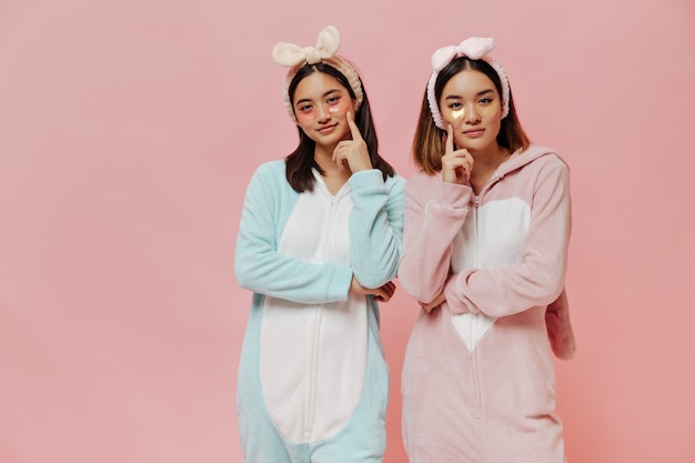 Free photo young attractive asian girls in cosmetic eyepatches look thoughtful at front, pose in pajamas on pink wall