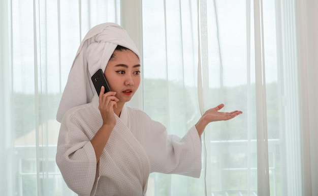 Young attractive asian woman in a bathrobe standing and talking on the phone at the living room
