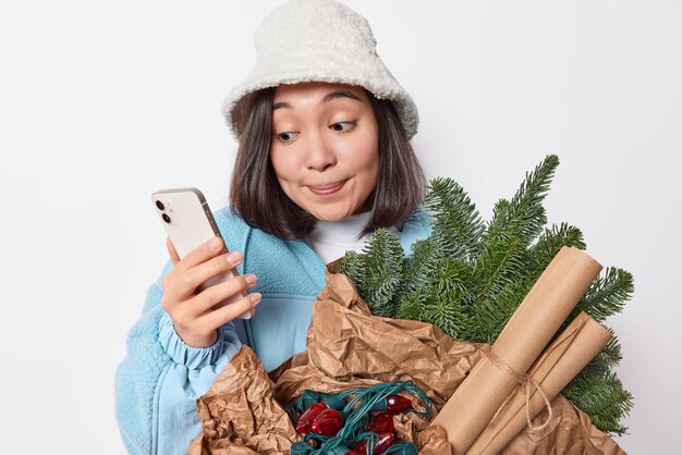 冬の服を着た若い魅力的なアジアの女性は、オンラインでチャットするために携帯電話を使用して、紙の花輪に包まれた緑のトウヒの枝を運び、新年のパーティーへの招待状を友達に送ります