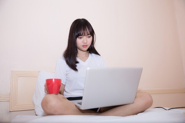 Молодая привлекательная азиатская женщина используя портативный компьютер пока лежащ на кровати в вскользь одеждах