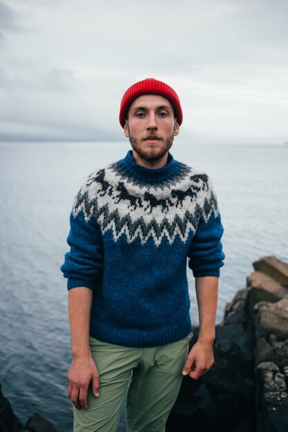 Молодой привлекательный бородатый миллениал в красной шляпе рыбака или моряка и синем свитере с традиционным исландским орнаментом