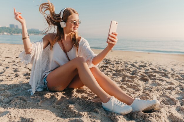 Young attractive blond smiling woman taking selfie photo on phone on vacation sitting on beach