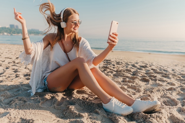 해변에 앉아 휴가에 전화 selfie 사진을 복용 젊은 매력적인 금발 웃는 여자