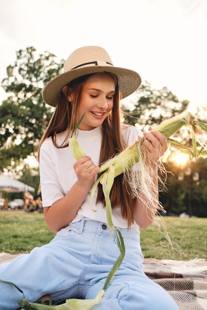 Young attractive brown haired girl in straw hat happily cleaning corn on picnic in city park