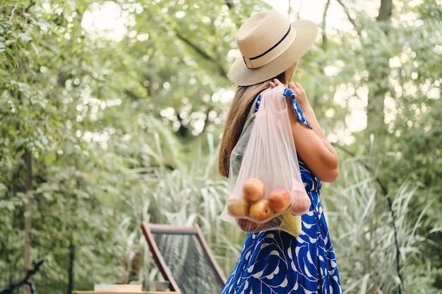 美しい公園で後ろから立っている間肩に果物と野菜のエコバッグを保持しているドレスと帽子の若い魅力的な茶色の髪の女性