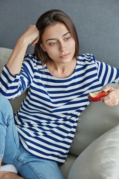 Young attractive brunette woman in casual striped sweater, eats pear, sits on sofa, has thoughtful expression, thinks about something