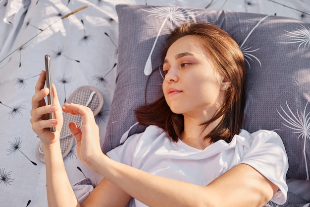 屋外のベッドの柔らかい枕の上に横たわって、携帯電話を使用して若い魅力的なブルネットの女性