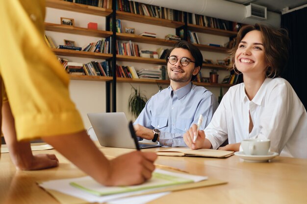 Young attractive business colleagues sitting at the desk with laptop happily looking at boss while working with papers in modern office