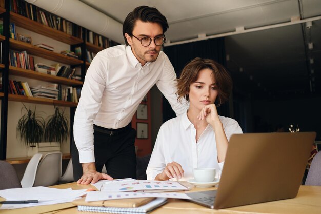 Young attractive business colleagues thoughtfully working on new project with laptop and papers in modern office