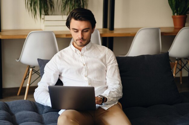 Молодой привлекательный бизнесмен сидит на диване и задумчиво работает на ноутбуке в современном коворкинге