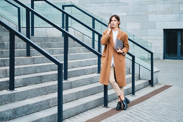 Young attractive businesswoman in coat with laptop confidently talking on cellphone on city street