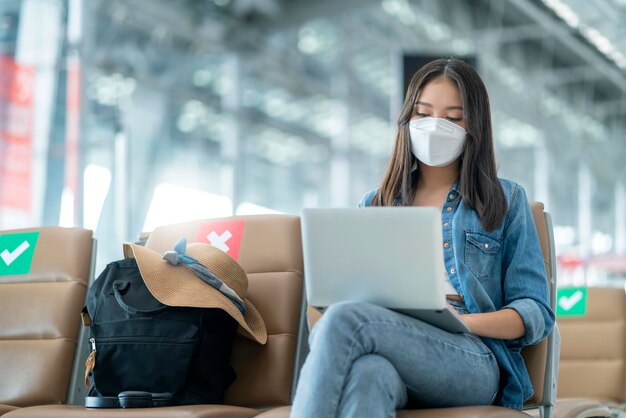 얼굴 마스크를 쓴 젊고 캐주얼한 아시아 여성 성인 여성은 공항 터미널 대기실에서 사회적 거리를 두고 앉아 노트북 기술을 사용하여 휴식을 취합니다.