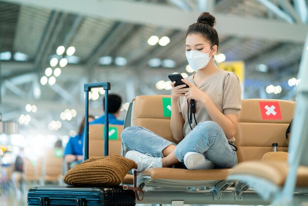 フェイスマスクを身に着けている若い魅力的なカジュアルなアジアの女性の大人の女性は、空港ターミナルの待合エリアで社会的な距離で座るスマートフォンソーシャルメディア技術を使用してリラックスして座ってください