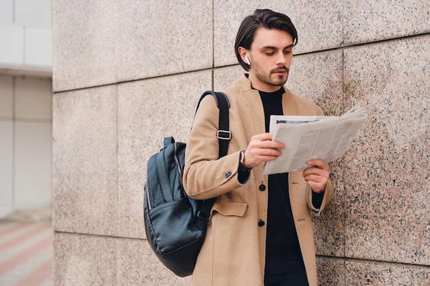 Молодой привлекательный случайный мужчина в бежевом пальто с рюкзаком задумчиво читает газету на открытом воздухе