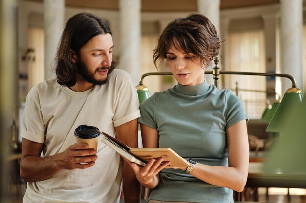 Young attractive casual students dreamily studying together with coffee and book in library of university