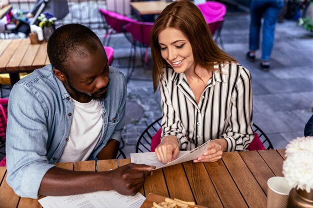 Young attractive caucasian girl and african boy are learning menu before ordering food