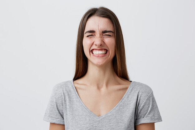 Young attractive cheerful caucasian student girl with dark long hair in casual gray t-shirt laughing brightly wit closed eyes on funny joke of friend in university, being happy and carefree.