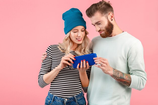 Young attractive couple listening to music on wireless speaker wearing cool stylish outfit smiling happy positive mood posing on pink background 