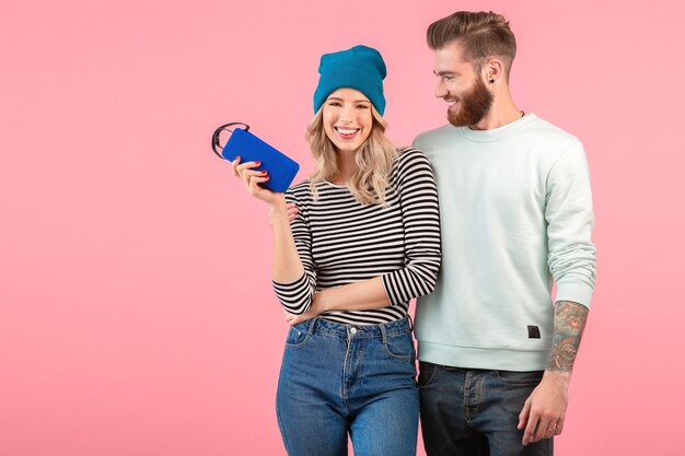 Young attractive couple listening to music on wireless speaker wearing cool stylish outfit smiling happy positive mood posing on pink wall isolated