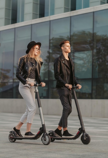 Young attractive couple on trendy scooters are riding in the city, near big glass building.