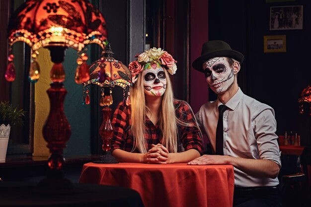 メキシコ料理のレストランでデート中に注文を待っているアンデッドの化粧をした若い魅力的なカップル。ハロウィーンとムエルトスのコンセプト。